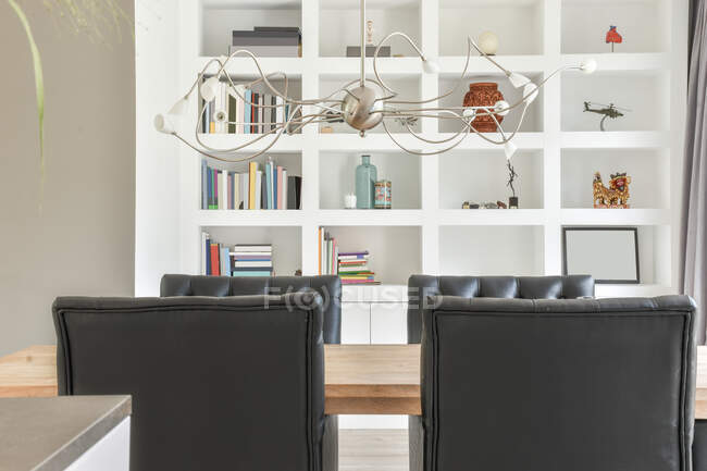 Порожній дерев'яний стіл і чорні шкіряні стільці розміщені в стильній кімнаті проти білих полиць з книгами та декоративними елементами — стокове фото