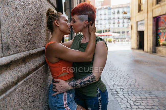 Vista lateral do jovem casal lésbico na moda com olhos fechados e tatuagem abraçando no momento do beijo na cidade — Fotografia de Stock