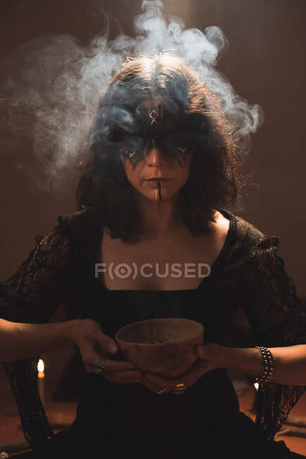 Bruxa de vestido preto e com rosto pintado de pé com tigela no quarto com vapor durante o ritual espiritual — Fotografia de Stock