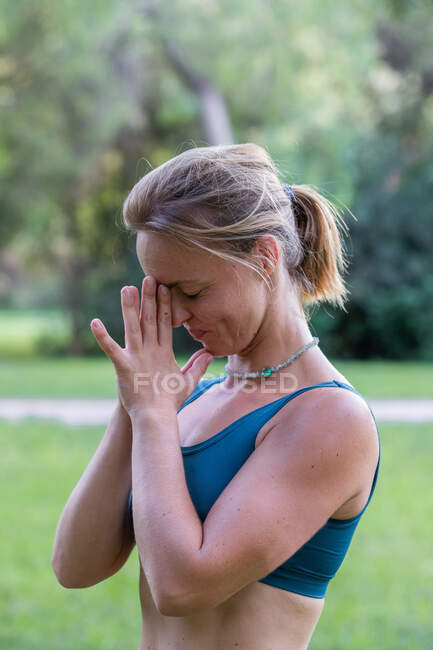 Vista lateral de la joven hembra en ropa deportiva de pie con las manos namaste mientras practica yoga en el parque verde durante el día - foto de stock