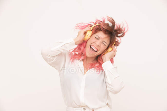 Вид спереду щасливої жінки з заплутаним і рожевим волоссям у білій блузці, що стоїть з закритими очима і слухає музику в навушниках на світлому фоні — стокове фото