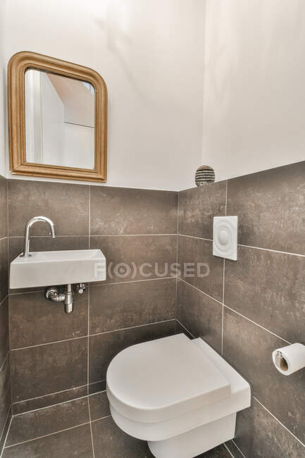 Современная ванная комната с зеркалом над умывальником против унитаза и бумажный рулон на серой керамической стене в доме — стоковое фото