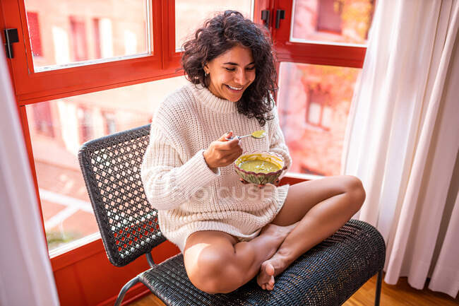 From above full body of barefoot latin female sitting with crossed legs ad eyes closed on chair and eating soup from bowl — Stock Photo