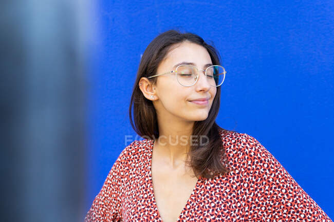 Щаслива жінка в стильній червоній сукні, що стоїть на синій стіні вдень з закритими очима — стокове фото