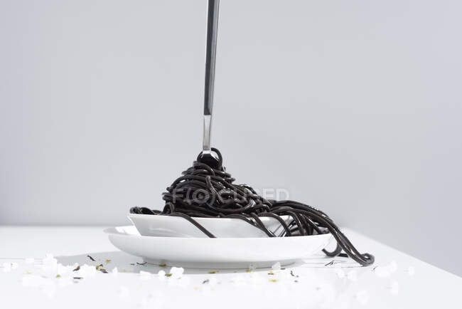Stainless fork in full bowl of black spaghetti with cuttlefish ink on white table with thin blooms in studio on gray background — Stock Photo