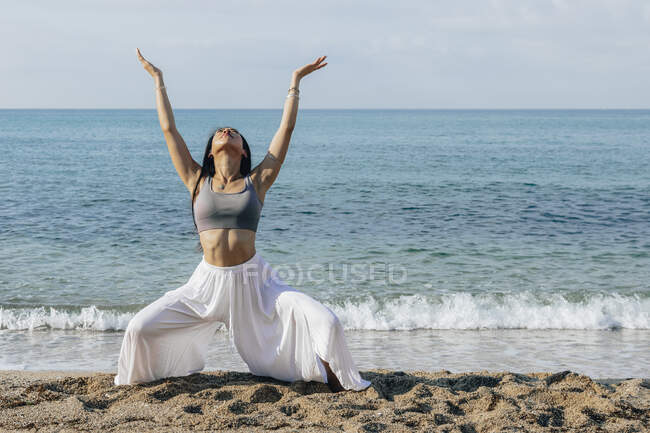 Молодая этническая женщина с поднятыми руками приседает на песчаном побережье, практикуя йогу против моря на солнце — стоковое фото