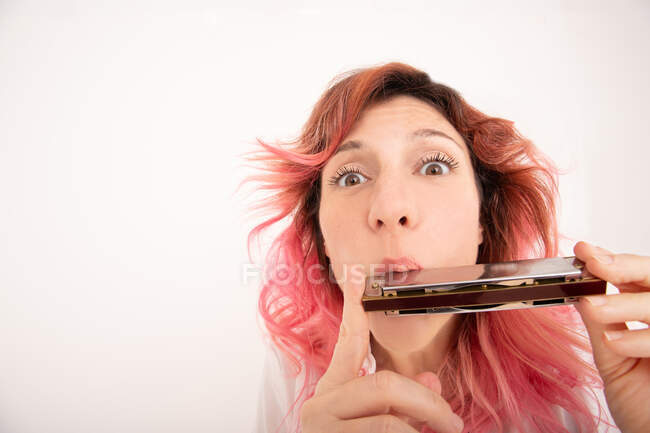 Musicista donna con i capelli rosa che suona armonica e guardando la fotocamera sullo sfondo chiaro in studio — Foto stock