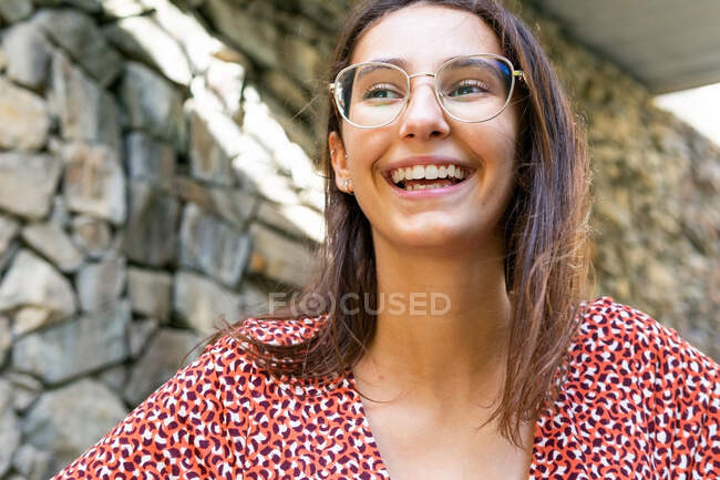 Joyeux jeune femme en robe rouge debout riant contre le mur de pierre dans la journée — Photo de stock