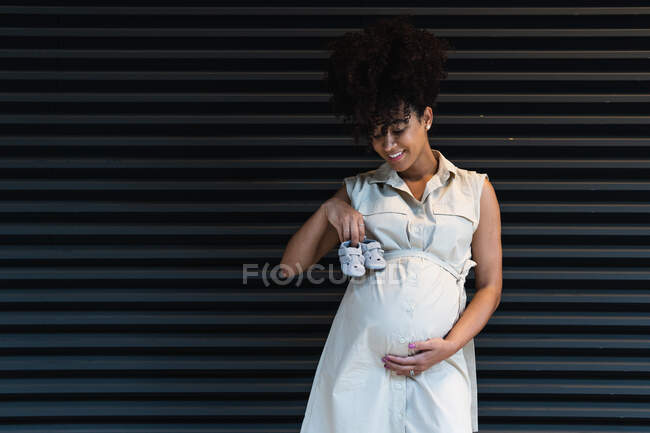Positive Afroamerikanerin mit lockigem Haar erwartet Geburt und zeigt Babybooties und blickt nach unten — Stockfoto