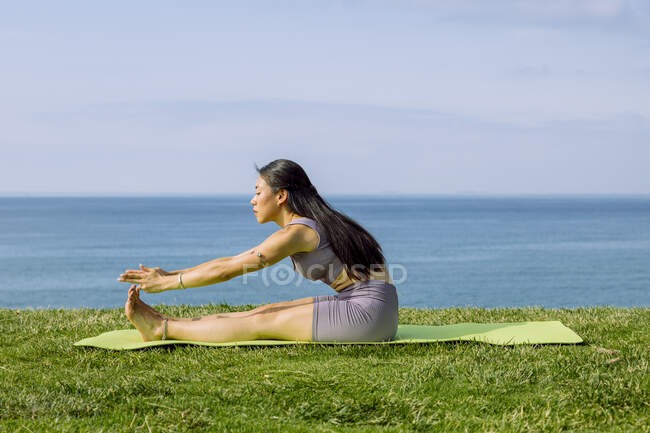 Вид сбоку на молодую этническую женщину, растягивающую ноги и спину, сидя в позе Пасхимоттанасаны на берегу океана — стоковое фото
