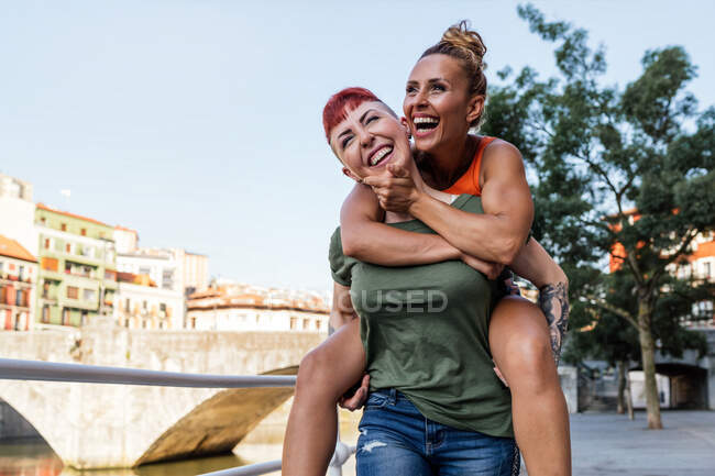 Positive lesbian female giving cheerful beloved piggyback ride while having fun against bridge and urban buildings — Stock Photo