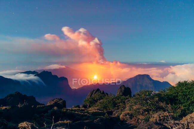 Paisaje nocturno con un volcán en erupción en el fondo y un mar de nubes que cubren las montañas desde una montaña vegetada y rocosa. Cumbre Vieja erupción volcánica en La Palma Islas Canarias, España, 2021 - foto de stock