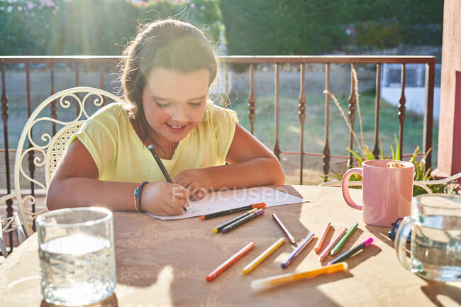 Ragazza positiva con i capelli scuri in abiti casual seduta a tavola con pennarelli e disegno su carta in terrazza nella giornata di sole — Foto stock