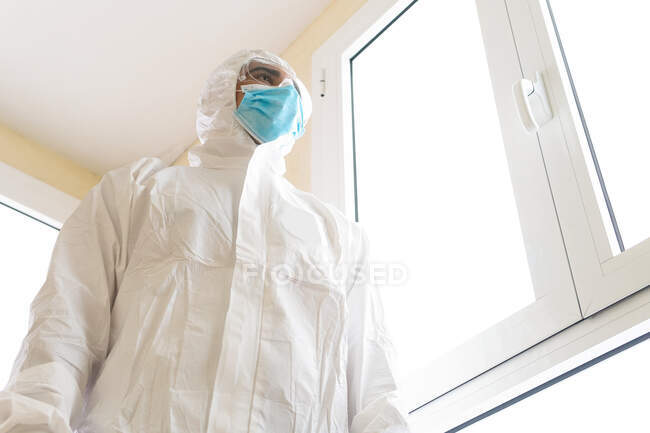 Снизу взрослый мужчина-медик в индивидуальном защитном снаряжении с очками и стерильной маской, смотрящий в окно в больнице — стоковое фото
