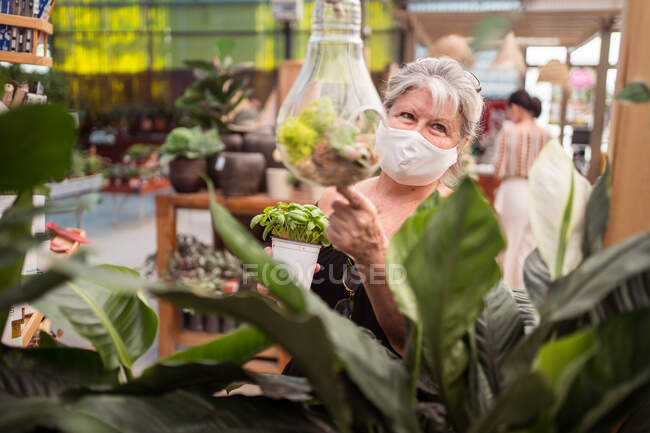 Comprador feminino de meia idade em máscara facial de pano olhando para longe enquanto apontando para a planta em florário no centro do jardim — Fotografia de Stock