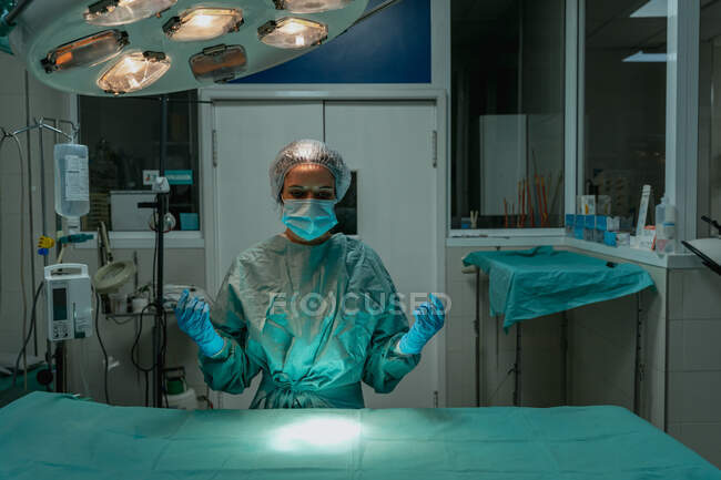 Jeune vétérinaire femelle en uniforme stérile et masque facial regardant vers le bas contre la table avec textile médical sous lampe à l'hôpital — Photo de stock