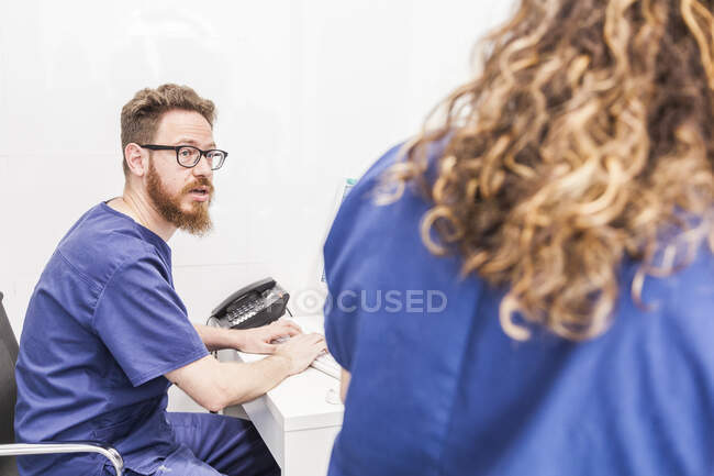 Medico maschio barbuto in uniforme e occhiali che digita sulla tastiera mentre lavora con il medico delle colture in ospedale — Foto stock