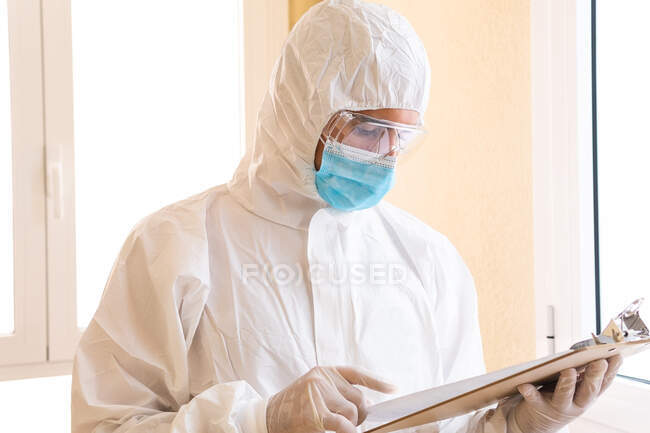 Médico varón adulto en equipo de protección personal papel de lectura en portapapeles durante la pandemia de COVID 19 en el hospital - foto de stock