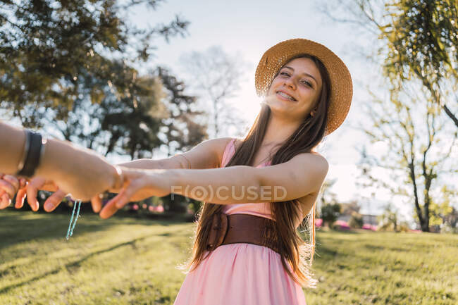 Улыбающаяся девушка-подросток в сарафане и соломенной шляпе держит анонимного партнера по урожаю вручную, глядя в камеру в парке — стоковое фото
