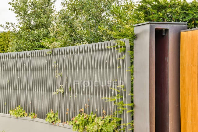 Творческий металлический забор на заднем дворе современного жилого дома с пышными зелеными деревьями и растениями в солнечный день — стоковое фото