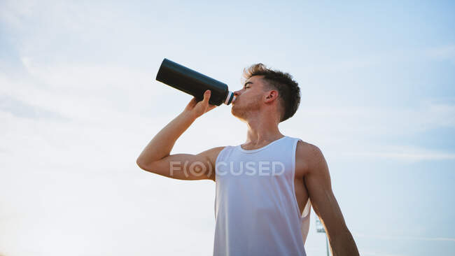 Giovane atleta di sesso maschile in abbigliamento sportivo con gli occhi chiusi bere acqua dalla bottiglia sotto il cielo nuvoloso — Foto stock