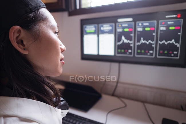 Вид сбоку концентрированной азиатской женщины, работающей на компьютере с графиками, показывающими динамику изменения стоимости криптовалют на удобном рабочем месте — стоковое фото