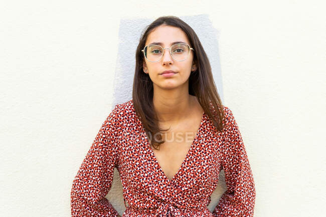 Femme sans émotion dans une robe élégante et des lunettes debout près du mur léger et regardant la caméra dans la journée — Photo de stock
