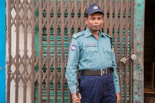 INDE, BANGLADESH - 6 DÉCEMBRE 2015 : Homme armé ethnique en uniforme de police et casquette debout près des portes métalliques du bâtiment endommagé et regardant la caméra — Photo de stock