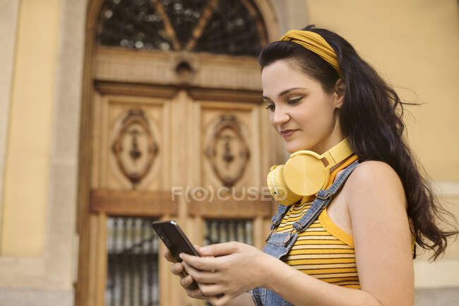 Вид збоку жінки в бездротових навушниках, що переглядають смартфон на вулиці, дивлячись на екран — стокове фото