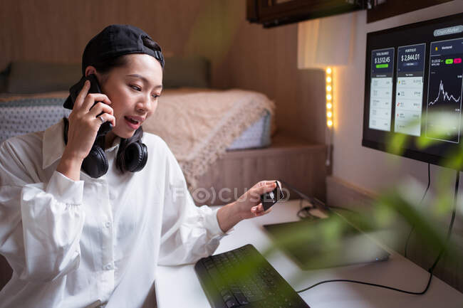 Surpreendida Asiática comerciante feminino falando no smartphone e olhando para o cartão de crédito enquanto trabalhava com criptomoeda estatística — Fotografia de Stock