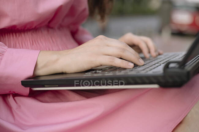 Cultivado irreconocible mujer de negocios en vestido sentado en el banco de trabajo en netbook en la calle de la ciudad - foto de stock