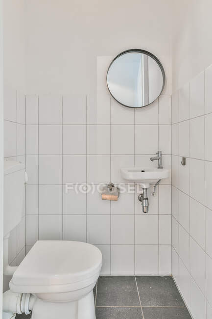 Cuarto de baño con inodoro contra lavabo bajo espejo redondo colgado en la pared de baldosas en la casa - foto de stock