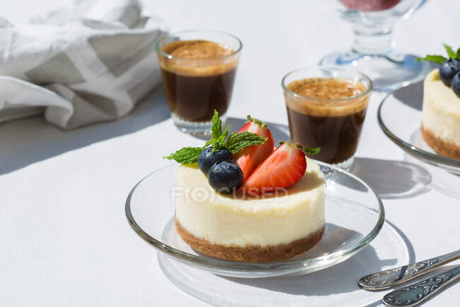 Sabrosos pasteles de queso con fresas frescas y arándanos bajo hojas de menta contra tragos de café expreso en la mesa en la cafetería - foto de stock