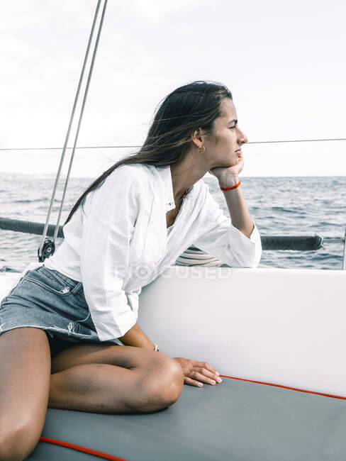 Adolescente contemplativa sentada con las piernas cruzadas en el banco de la lancha en el océano mientras mira hacia otro lado en Tenerife España - foto de stock