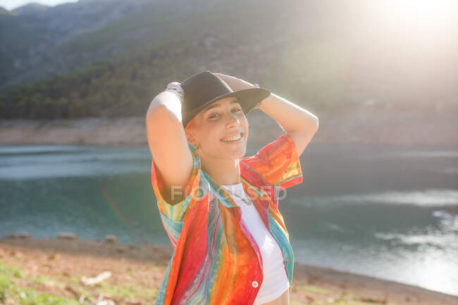 Mujer vista lateral en un lago, mirando una cámara con una mano sosteniendo un sombrero negro - foto de stock
