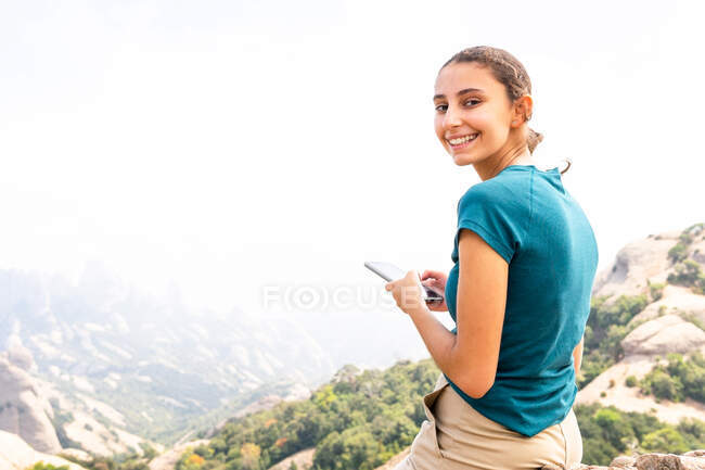 Vista laterale di messaggi turistici femminili positivi sul cellulare durante l'escursione nelle montagne di Montserrat in Spagna guardando la fotocamera — Foto stock