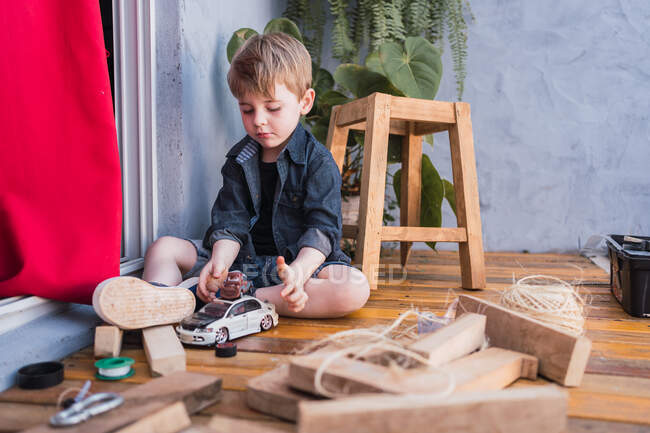 Charming child grimacing while playing with toy automobiles between wooden pieces and handmade stool in daylight — Stock Photo