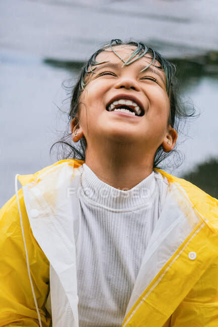 Content Asian child in slicker looking up while playing on rainy day — Stock Photo