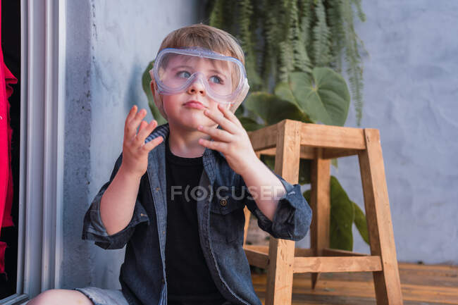 Contemplating child in safety glasses and denim shirt gesticulating while looking away against handmade stool in daytime — Stock Photo
