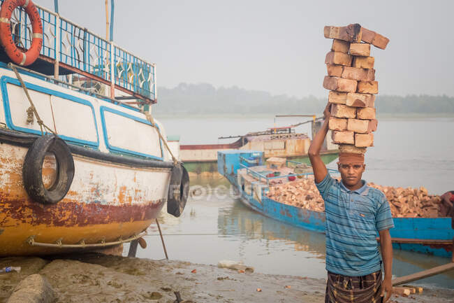 INDIA, BANGLADESH - 8 DE DICIEMBRE DE 2015: Joven hombre étnico con ropa sucia caminando con piedras de ladrillo sobre la cabeza cerca del río con barcos mirando a la cámara - foto de stock