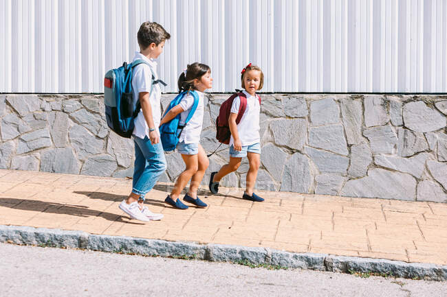 Вид сбоку школьника с рюкзаком, который разговаривает с подругами, прогуливаясь по плиточной тротуарной плитке на фоне каменной стены при солнечном свете — стоковое фото