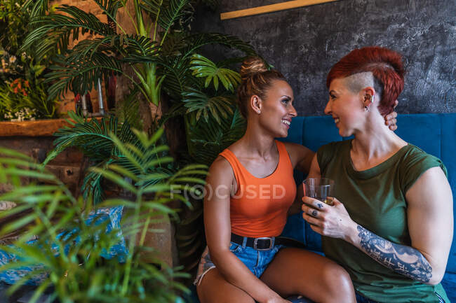 Содержание молодой татуированной женщины с ирокезом и напитком, обнимающей лесбиянку, глядя друг на друга на диване в доме — стоковое фото