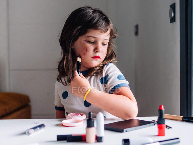 Niño con aplicador que compone la cara en la mesa con una variedad de productos cosméticos en casa - foto de stock