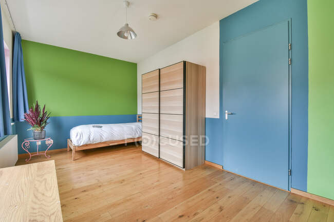 Ліжко розміщується в кутку біля шафи в мінімалістичній спальні з зеленими і синіми стінами і декоративною рослиною на скляному столі — стокове фото