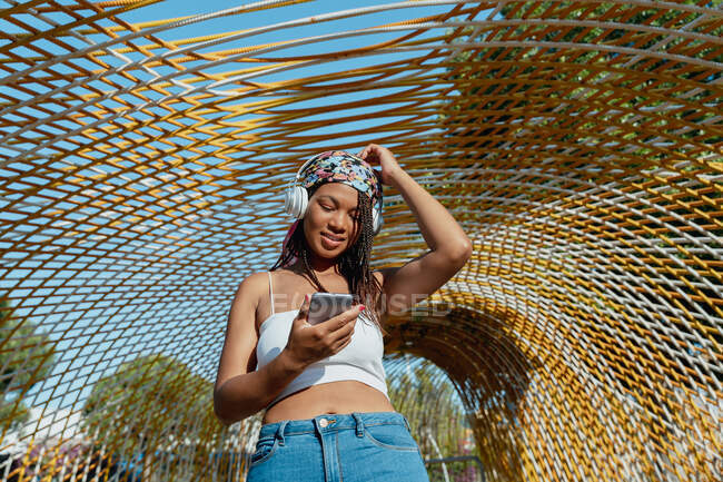 Позитивна молода етнічна жінка з мобільним телефоном слухає музику з бездротових навушників під час сміху в місті — стокове фото