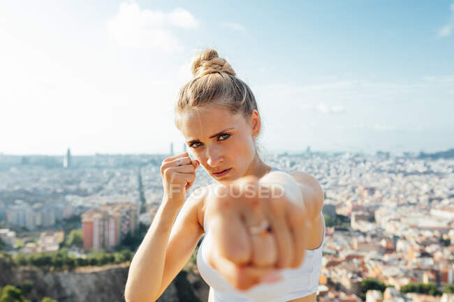Boxeadora gritando mientras muestra la técnica de golpear y mirando la cámara durante el entrenamiento en la ciudad soleada - foto de stock