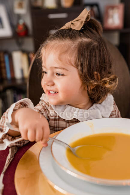 Bambino affascinante con fiocco su capelli castani e cucchiaio guardando lontano contro piatto di minestra di purè di zucca in casa — Foto stock