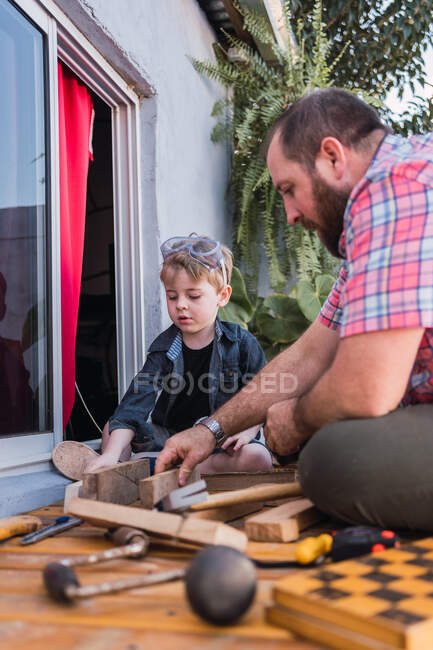 Im Erdgeschoss ein fröhlicher bärtiger Vater im karierten Hemd mit einem Jungen, der mit Holzklötzen arbeitet — Stockfoto