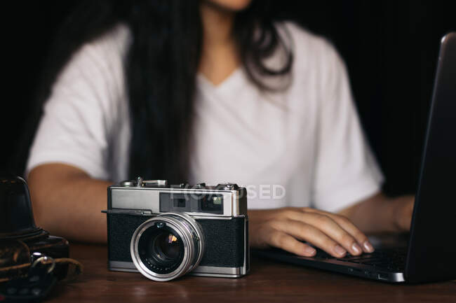 Crop freelancer femenina anónima trabajando en netbook contra la cámara de fotos vintage en la mesa sobre fondo negro - foto de stock