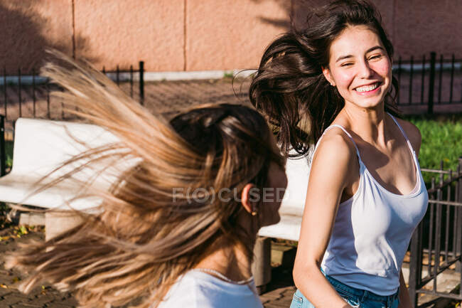 Веселые лучшие подруги с летящими каштановыми волосами смеются, проводя время в городе в солнечный день — стоковое фото